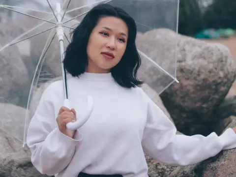 ErikaKim — Young woman in a white sweater holding a clear umbrella against a rocky outdoor background on a cloudy day.