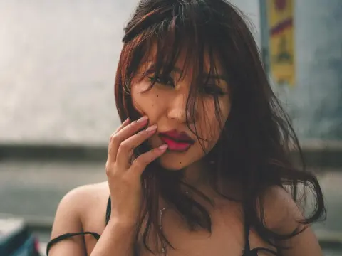 ErikaKim — ErikaKim poses with her hand touching her face, showing her long dark hair and red lips against a soft outdoor background.