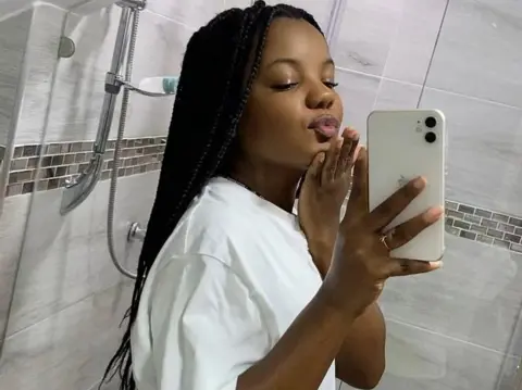 Young Black woman with long braids in a white t-shirt taking a playful mirror selfie in a modern tiled bathroom.