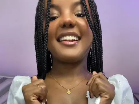 Smiling Black woman with long box braids wearing a white blouse and gold necklace in a bright indoor setting.