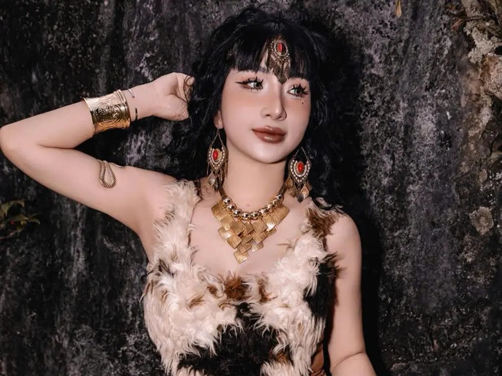 MirannLoves poses against a dark rock wall, wearing a brown furry top with long, wavy black hair and decorative face glitter.