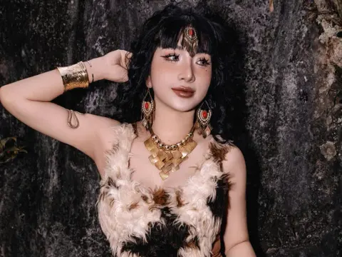 MirannLoves poses with one hand behind her head, wearing a furry white and brown top, heavy gold jewelry, and a red headpiece against a dark stone wall.