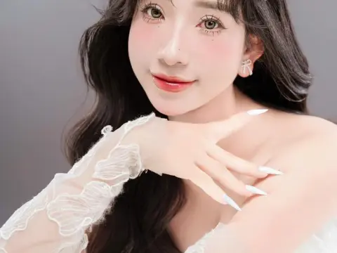 MirannLoves poses with her hand against her shoulder, wearing a white lace sleeve and white long nails against a plain grey background.