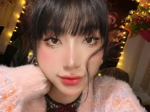 MirannLoves poses close-up with dark hair and bangs, wearing a pink fuzzy sweater and a black lace choker against a warm, lit background.