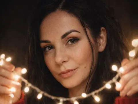 Smiling woman with dark hair holding glowing fairy lights in a cozy, warm indoor setting.
