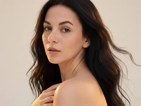 Portrait of a brunette woman with wavy hair looking serene against a neutral studio background.