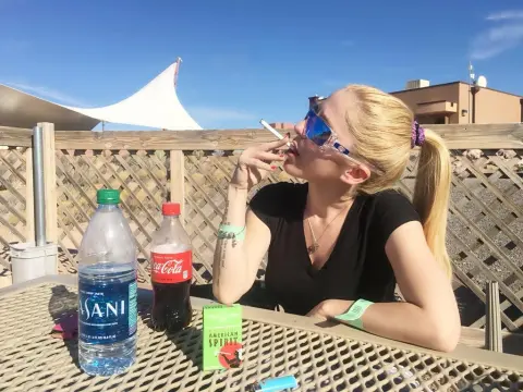 Exclusivegirl — Blonde woman in black top smoking a cigarette at an outdoor sunny patio table with drinks.