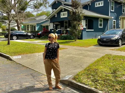 Exclusivegirl — Woman in black patterned t-shirt and khaki trousers standing on a sunny residential street by a blue house.