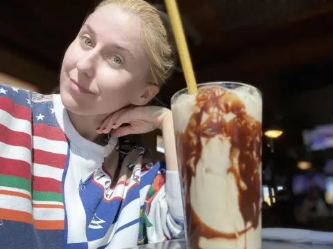 Exclusivegirl — Blonde woman in a colorful striped sweatshirt enjoying a chocolate milkshake in a cozy cafe setting.