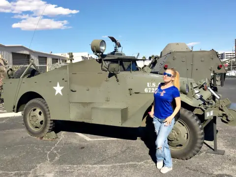 Exclusivegirl — Woman in blue t-shirt and sunglasses standing by a vintage olive green US military armored vehicle outdoors.