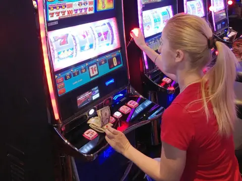 Exclusivegirl — Blonde woman in a red shirt playing a slot machine game in a brightly lit casino.