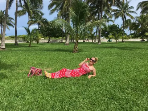Exclusivegirl — Relaxed woman in a red patterned sundress lounging on green grass under tropical palm trees.