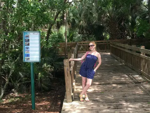 Exclusivegirl — Smiling woman in a blue polka dot romper standing on a wooden boardwalk path in a lush tropical garden.