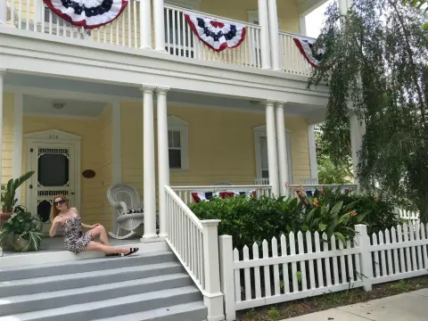 Exclusivegirl — Stylish woman in a patterned sundress relaxing on the porch of a festive yellow colonial-style house.