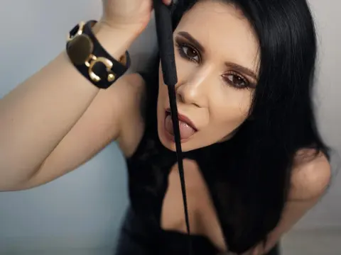 AlisiaChase — Glamorous woman with long black hair and gold bracelet holding a black whip in a moody studio setting.
