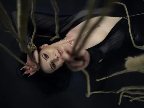 AlisiaChase — Elegant woman in a black dress lying amidst thick ropes in a moody, dark cinematic setting.