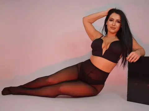 Smiling brunette woman in black lingerie and sheer tights posing against a soft pink studio background.