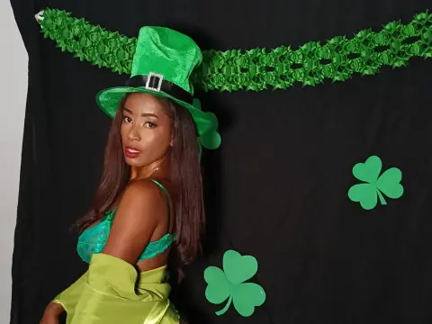BrendaRos wears a green leprechaun hat and green lace lingerie while posing against a black background with shamrock decorations.
