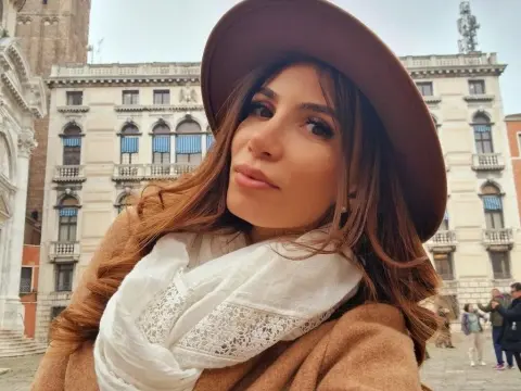 Smiling woman in a brown wool coat and white lace scarf wearing a wide-brimmed hat in a European city square.