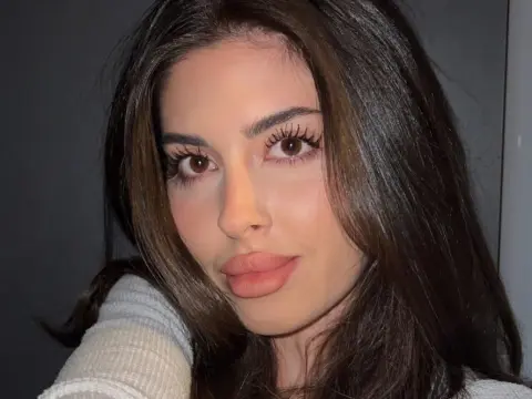EvaBerryy — EvaBerryy looking at the camera with long dark hair and heavy eyelashes while wearing a white ribbed top.