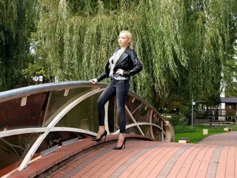 EmmaKyle — Blonde woman in a black leather jacket and jeans posing on a curved bridge amidst lush green willow trees.