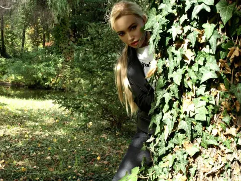 EmmaKyle — Blonde woman in a black leather outfit peeking through green ivy in a sunny garden setting.