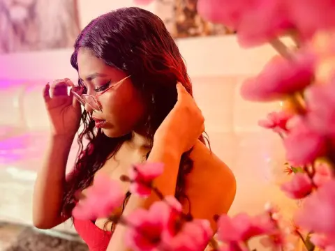 Young woman with wavy hair wearing glasses in a warm, dreamy setting surrounded by pink flowers.