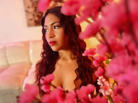 Chloe_Lombardi poses with long, wavy dark hair, looking off-camera behind bright pink flowers.