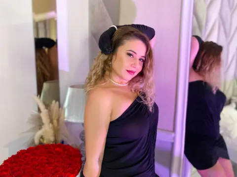Woman in a black one-shoulder top and headband posing near red roses in a soft pink lit room.