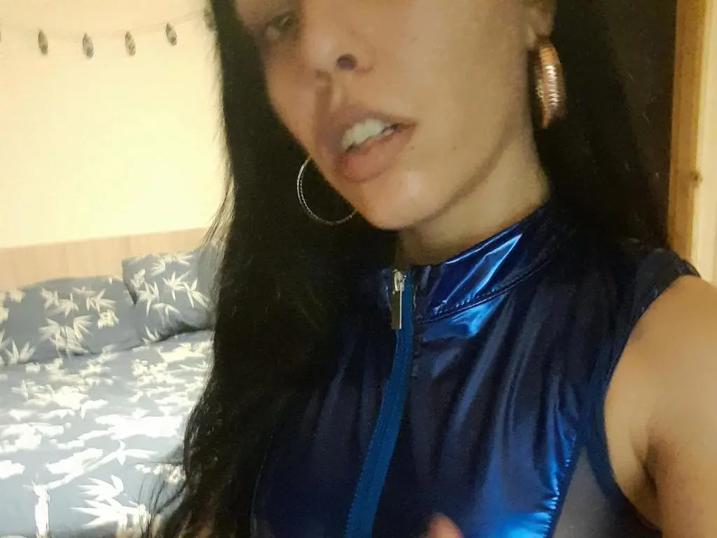 IconDomme808 poses in a blue shiny zip-up top with long dark hair and gold hoop earrings.