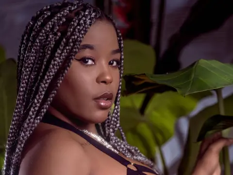 AshantyHopsom with long grey braids poses amidst large green tropical leaves in a dark setting.
