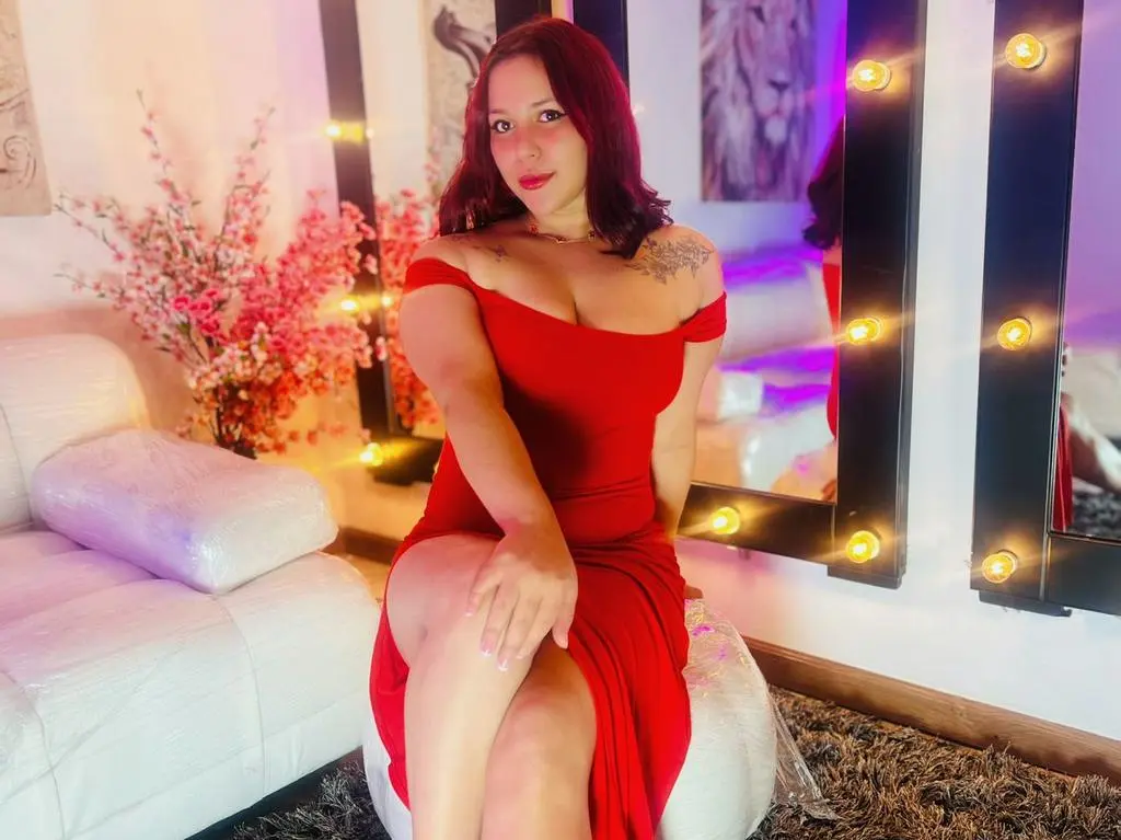ivy_browm wearing a red off-the-shoulder dress, sitting on a white stool in front of a lit vanity mirror with pink flowers behind her.