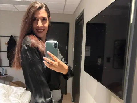 Smiling woman in a black silk blouse taking a mirror selfie in a modern bedroom setting.