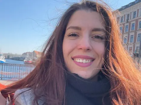 Smiling woman with long brown hair wearing a black turtleneck in a sunny outdoor harbor setting.