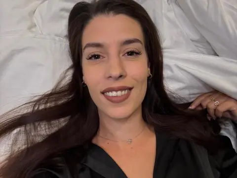Smiling young woman with long brown hair wearing a black silk pajama top while relaxing on white bedding.