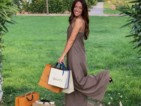 SpoiledCarlota — Smiling woman in a brown maxi dress carrying Gucci shopping bags in a sunny green garden.