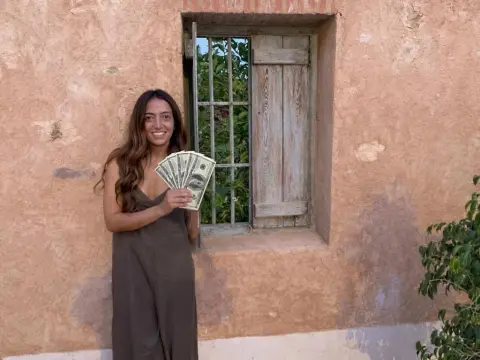 SpoiledCarlota — Smiling woman in a brown dress holding US dollar bills against a rustic textured wall background.