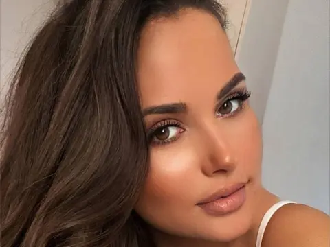 Close-up portrait of a beautiful brunette woman with glowing makeup and brown eyes in a bright indoor setting.