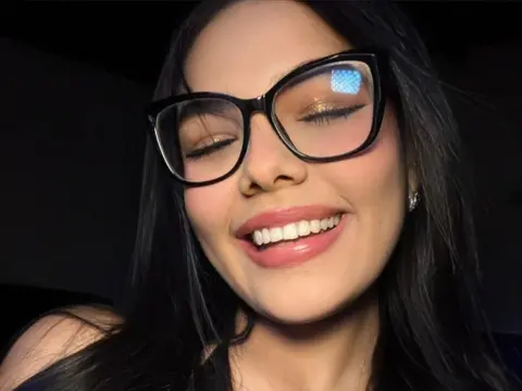 Smiling young woman with long dark hair wearing stylish black glasses in a moody, low-light setting.