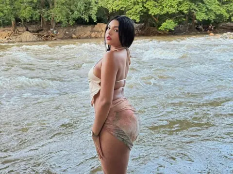 Young woman in a beige bikini and patterned sarong posing by a rushing river in a lush forest setting.