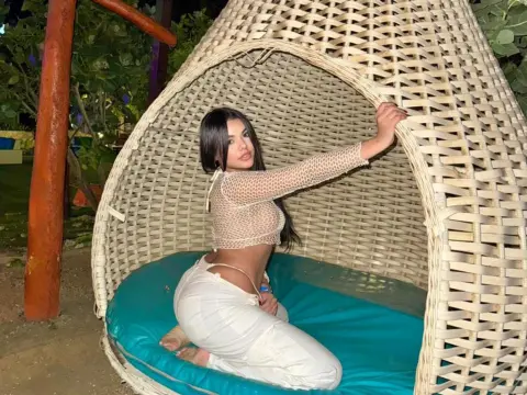 Young woman in a white mesh crop top and trousers relaxing in a wicker hanging egg chair at night.