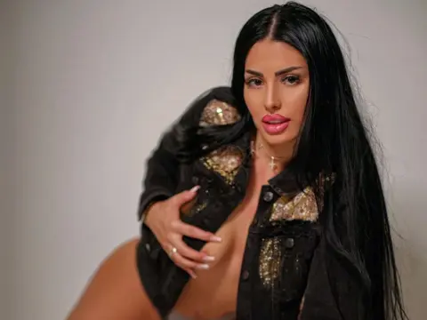 CindyTailX poses with long black hair, wearing an open black denim jacket that shows her bare chest and cleavage against a plain background.