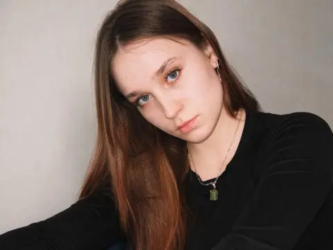 Young woman with long brown hair wearing a black long-sleeve shirt and green crystal necklace in a soft indoor setting.