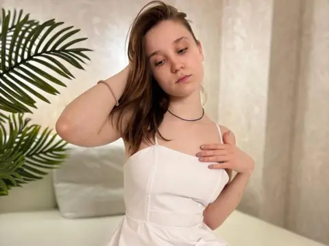 Young woman in a white spaghetti strap dress posing thoughtfully next to a green palm plant in a bright room.