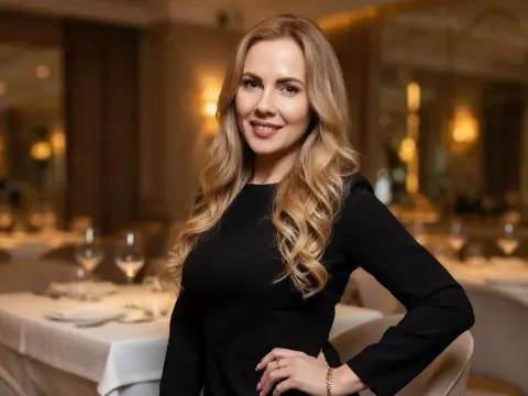 Janie_Kay is a smiling blonde woman wearing a tight black long-sleeved dress, posing in a fancy restaurant setting.