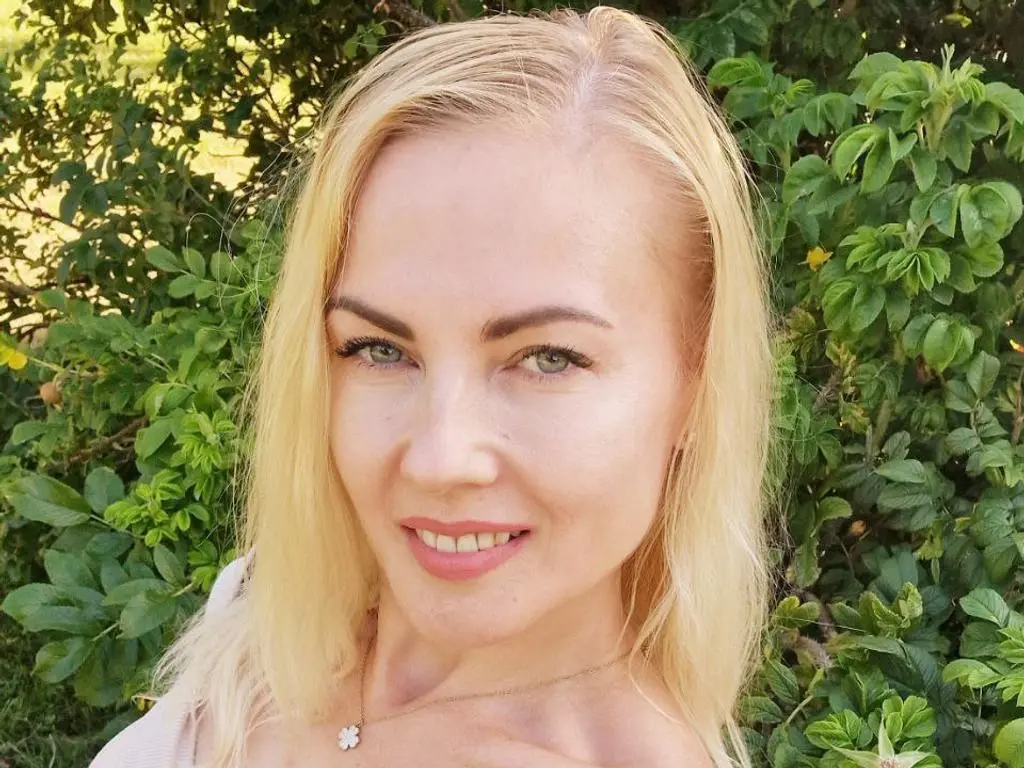 Janie_Kay poses with a smile, her blonde hair falling over her bare shoulders against a forest backdrop.