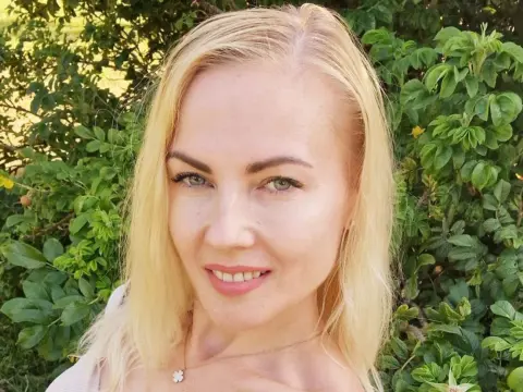 Janie_Kay is a blonde woman smiling in front of green leafy bushes.