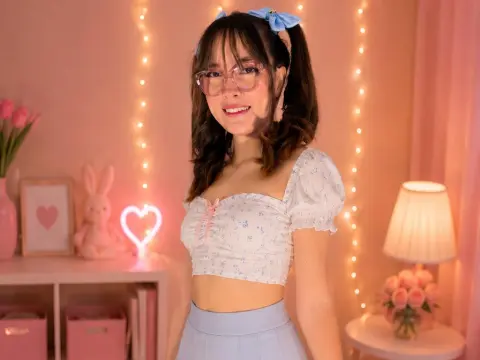 Smiling young woman in a white floral crop top and blue skirt in a cozy, pink aesthetic bedroom setting.