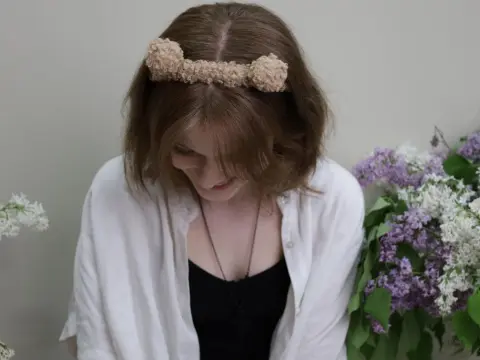 YukiPage — YukiPage looks down with a smile, wearing a white shirt and a fuzzy headband while sitting near purple lilacs.
