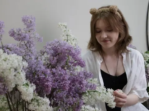 YukiPage wears a white shirt and bear ears while smiling and smelling a large bouquet of purple and white lilacs.